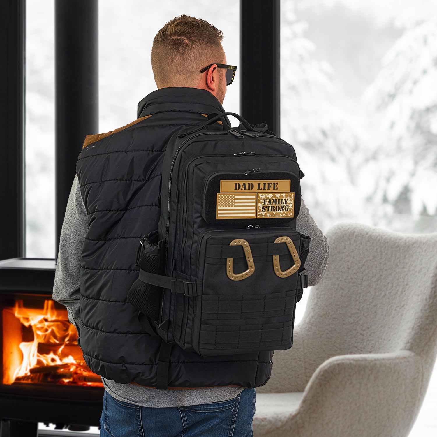 Man wearing a black dad diaper bag backpack with 'Dad Life' and 'Family Strong' patches by a fireplace.