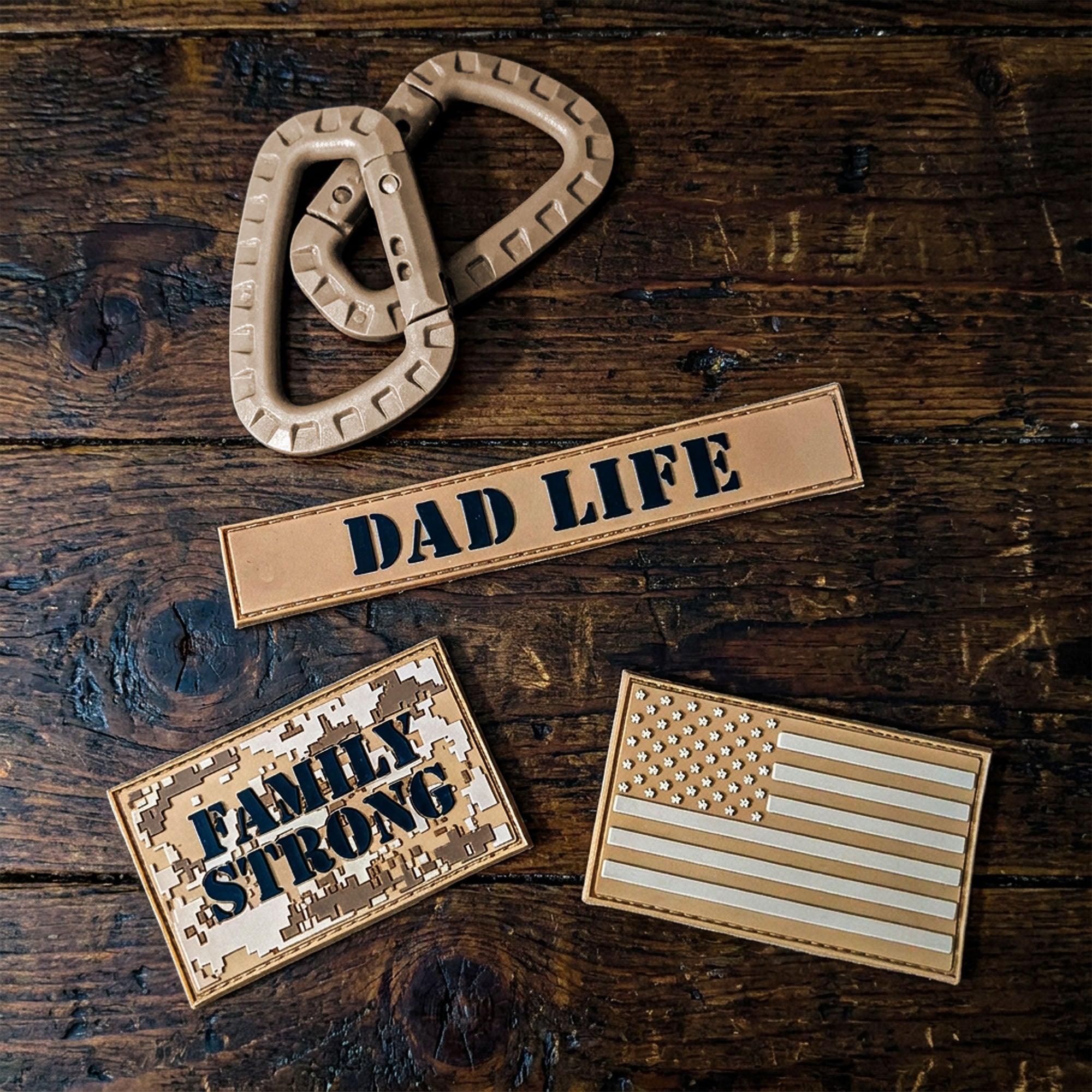 Three tan dad patches displayed on a dark rustic wooden surface: a carabiner-shaped patch with no text, a rectangular patch reading "DAD LIFE," and a rectangular patch reading "FAMILY STRONG" alongside an American flag design.