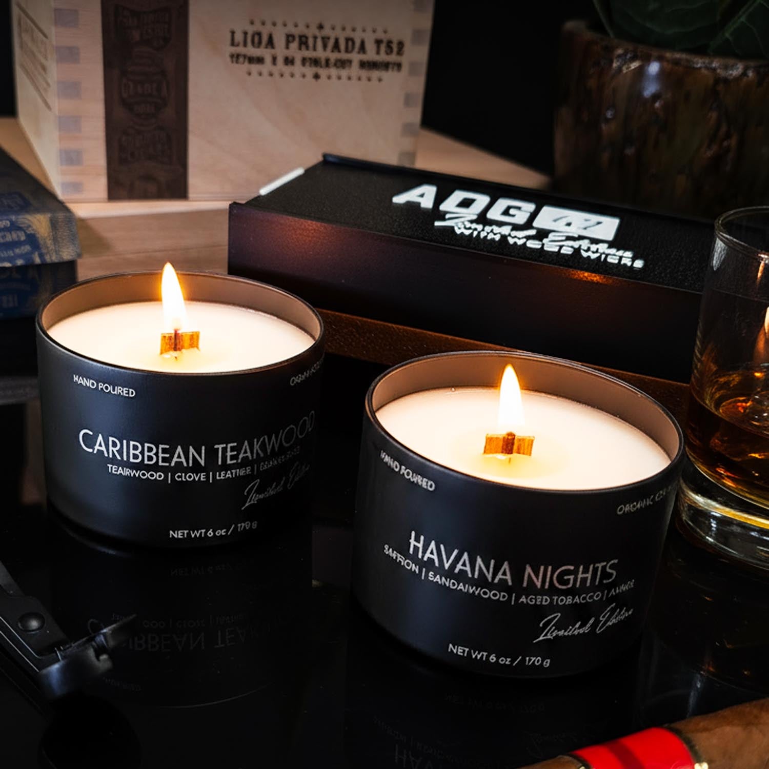 Two lit candles labeled 'Caribbean Teakwood' and 'Havana Nights' on a table with a glass of whiskey and a box in the background.