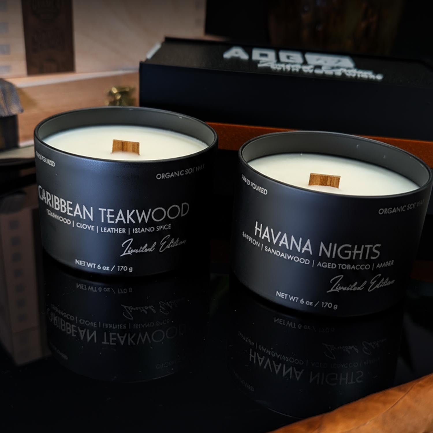 Two candles in black tins labeled 'Caribbean Teakwood' and 'Havana Nights' on a reflective surface.