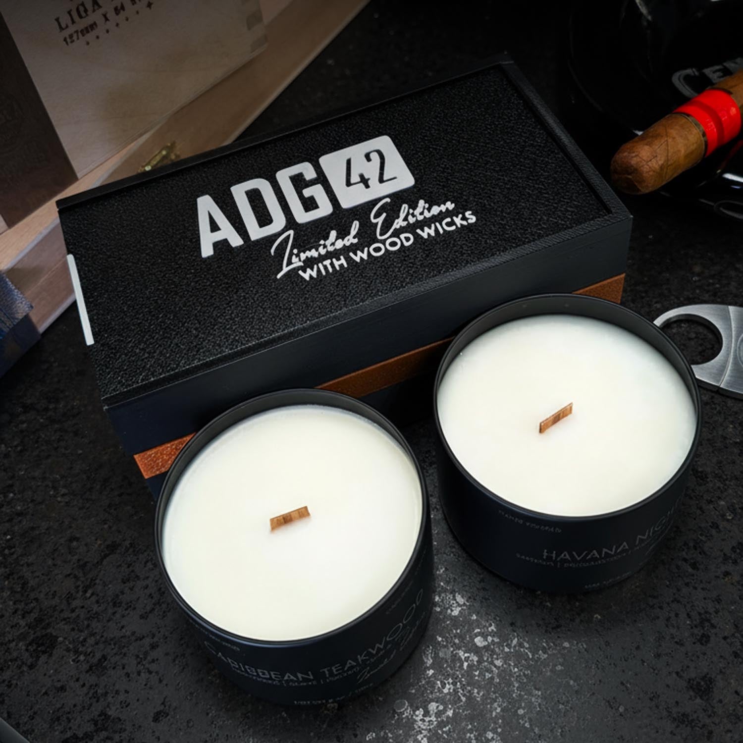 Two candles with wood wicks in black containers next to a branded box on a dark surface.