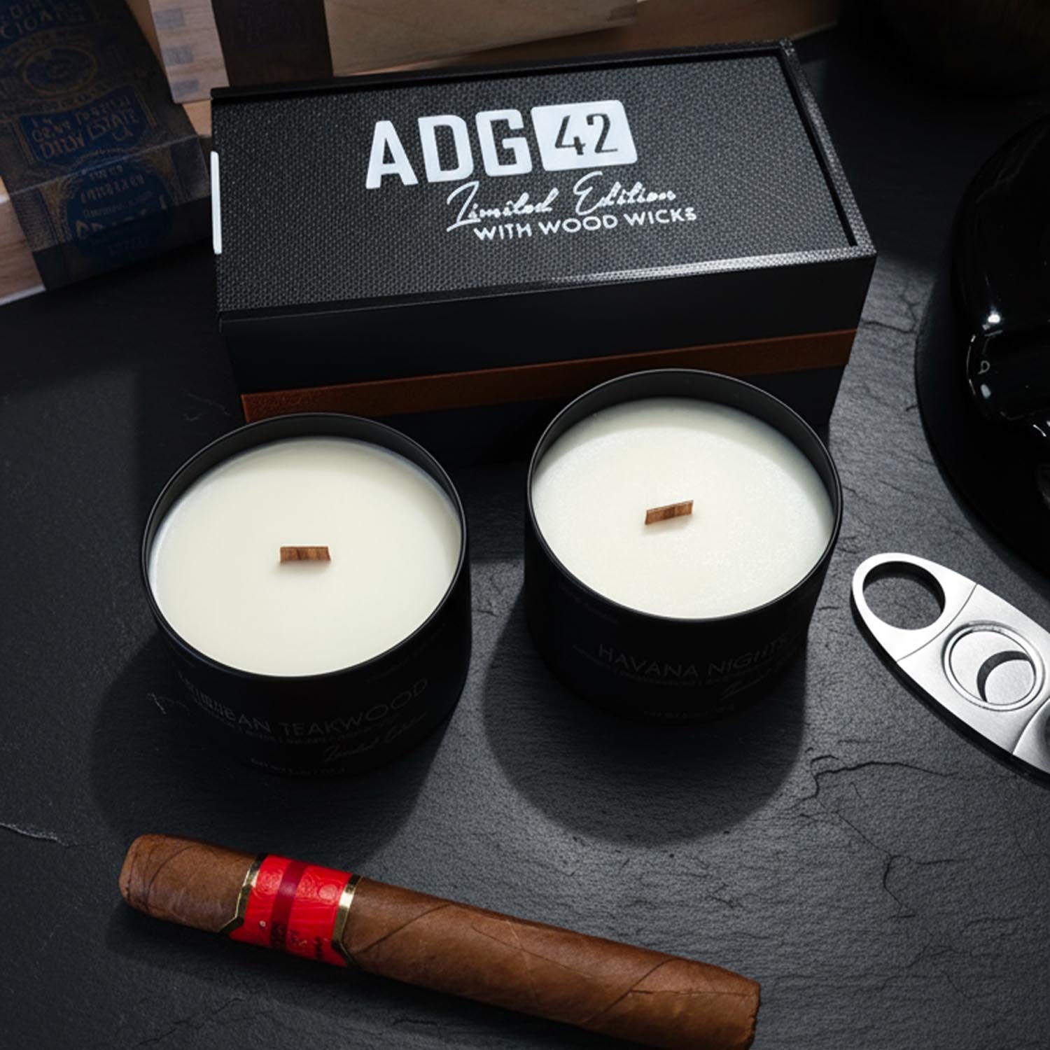 Two black candles for men with wood wicks on a dark surface, accompanied by a cigar and a box labeled 'ADG 42'.